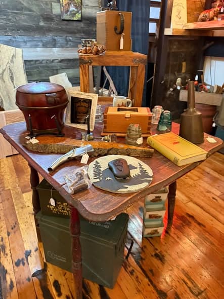 A rustic display table with military ammo cans, vintage tools, and collectibles on hardwood floors
